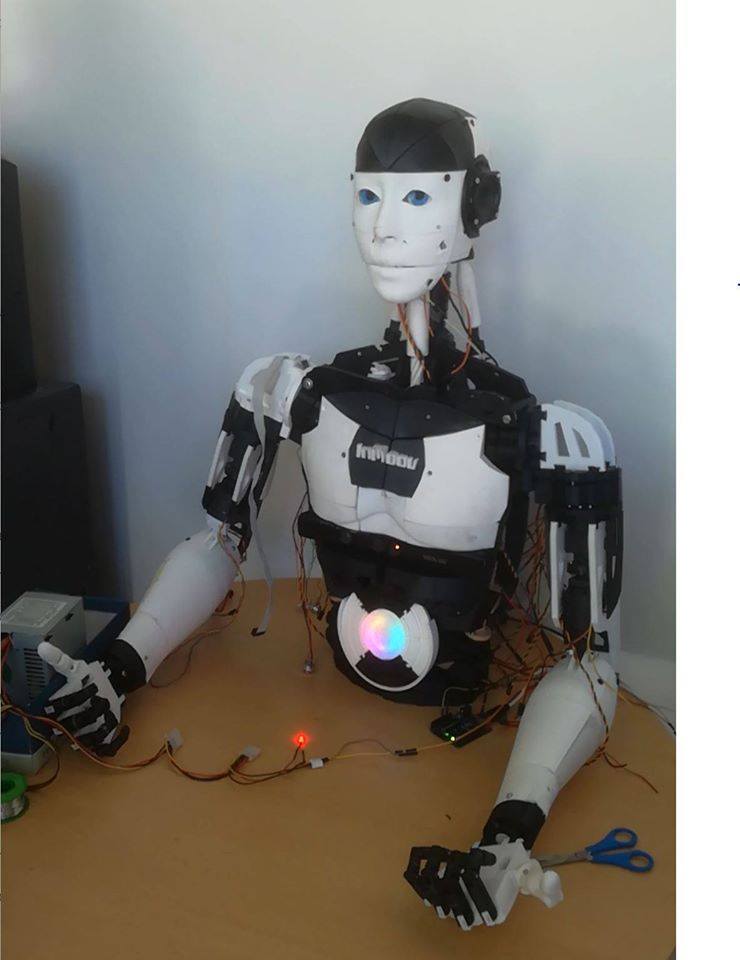 The InMoov robot seated at a desk with both arms wired up and an RGB light glowing in its chest.