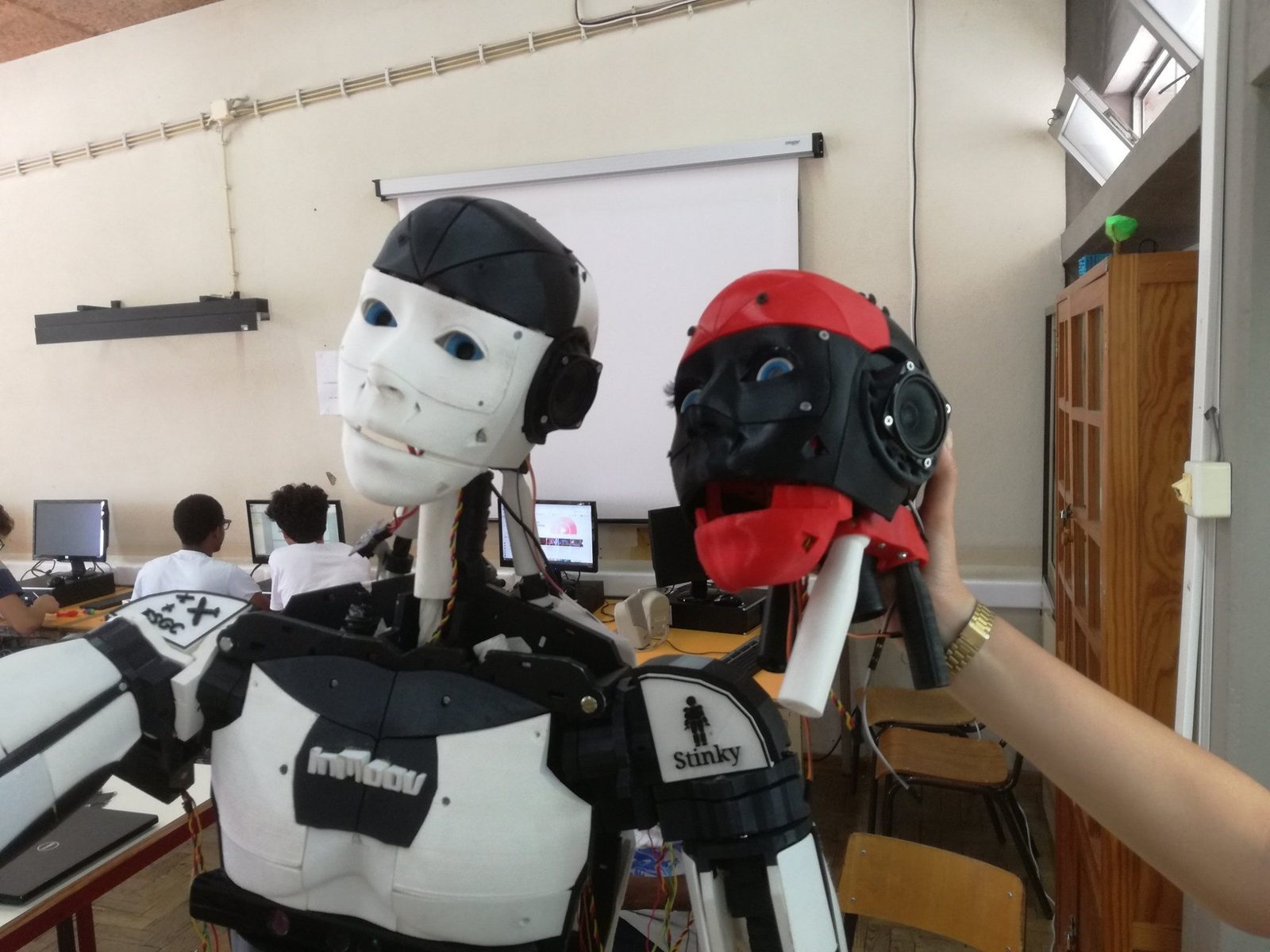 The assembled InMoov torso in a computer lab while a hand holds a second red and black head next to it.