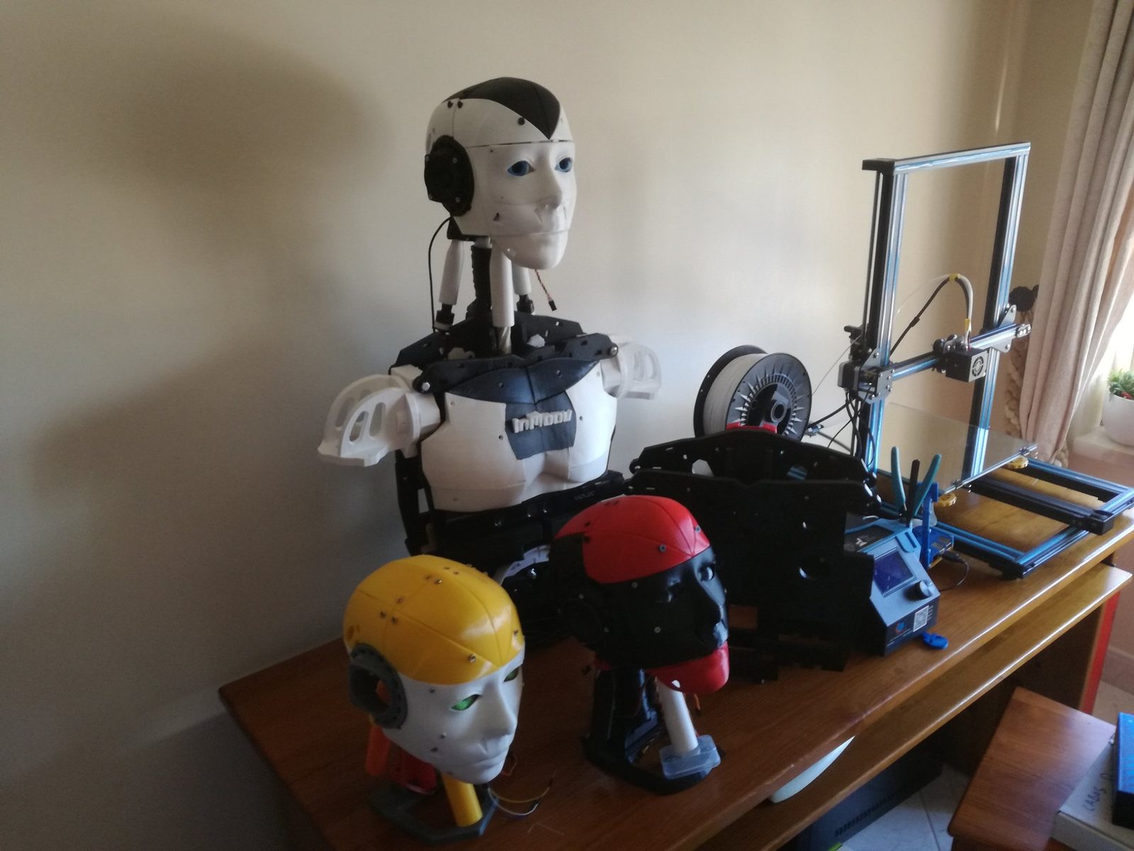 The robot torso on a table beside a running 3D printer and two spare printed heads, one yellow and one red and black.