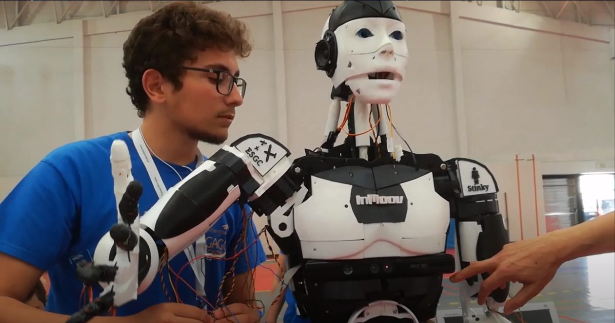 A club member stands beside the completed InMoov robot while a visitor reaches out to touch its arm.