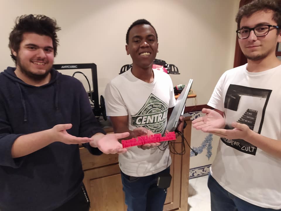 Three team members presenting a red 3D-printed trophy to the camera, with the 3D printer in the background.
